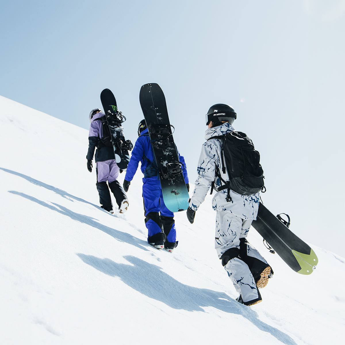 Three snowboarders, dressed in winter gear including helmets, goggles, and backpacks, are walking up a snowy mountain slope while carrying their snowboards. The image captures their journey against a bright sky, with crisp shadows on the pristine snow.